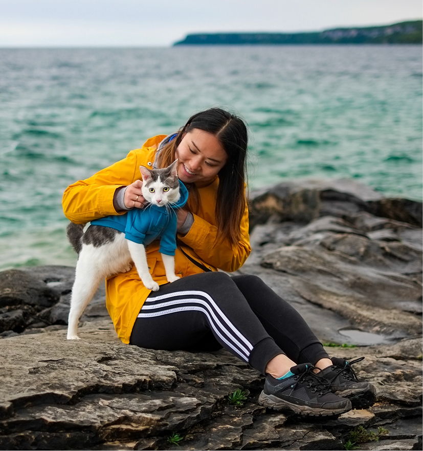 Person in yellow jacket cuddling a cat on rocky shore by the water.