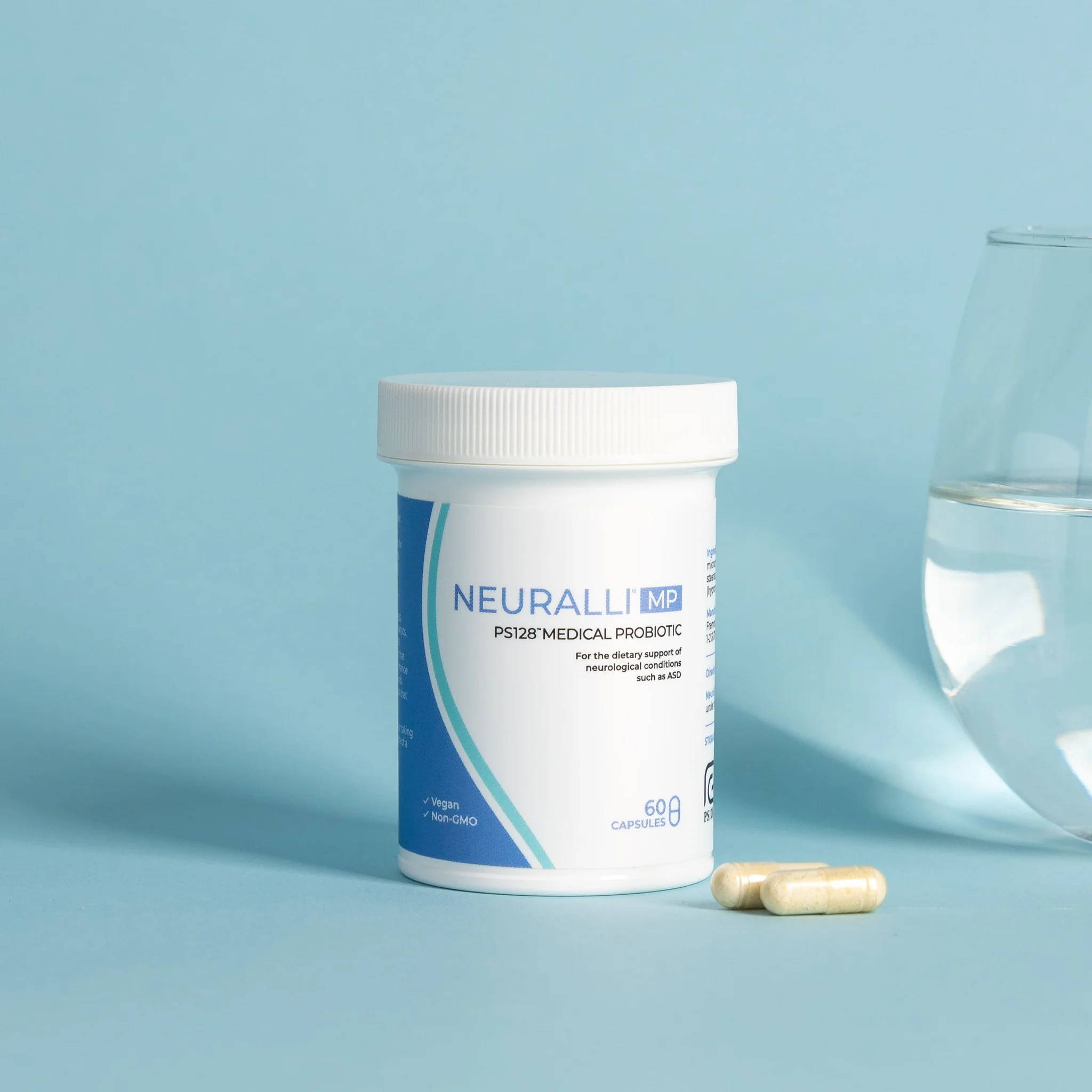 A bottle of Neuralli MP probiotic medical food against a blue background, with two capsules lying next to it. To the far right is a glass of water.