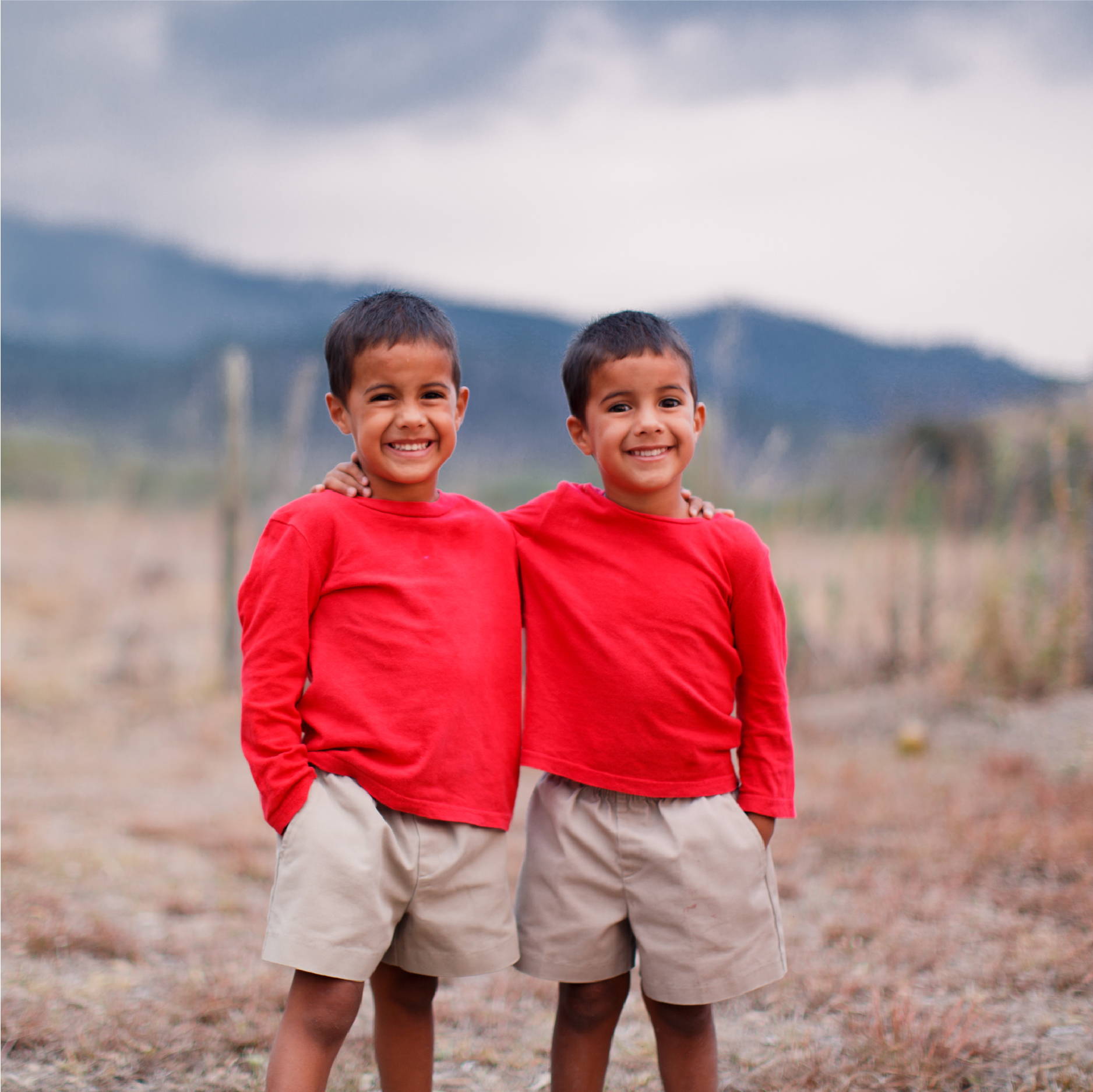 two young Honduran boys wear khaki shorts and red shirts as they stand and smile in front of a mountain range in Honduras. 