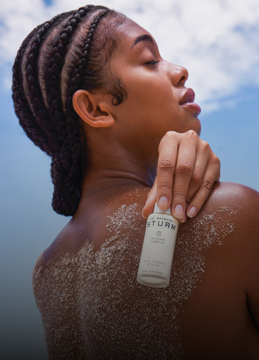 Person with braids applying skincare product on their back outdoors.