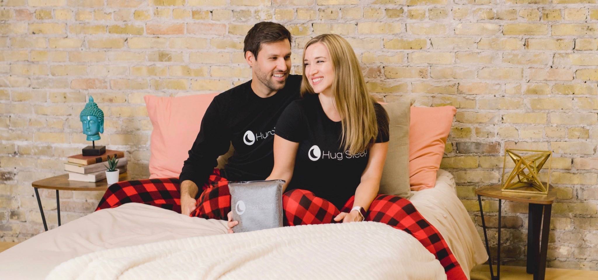 A photo of a man and woman sitting on a bed holding a Sleep Pod in its packaging 