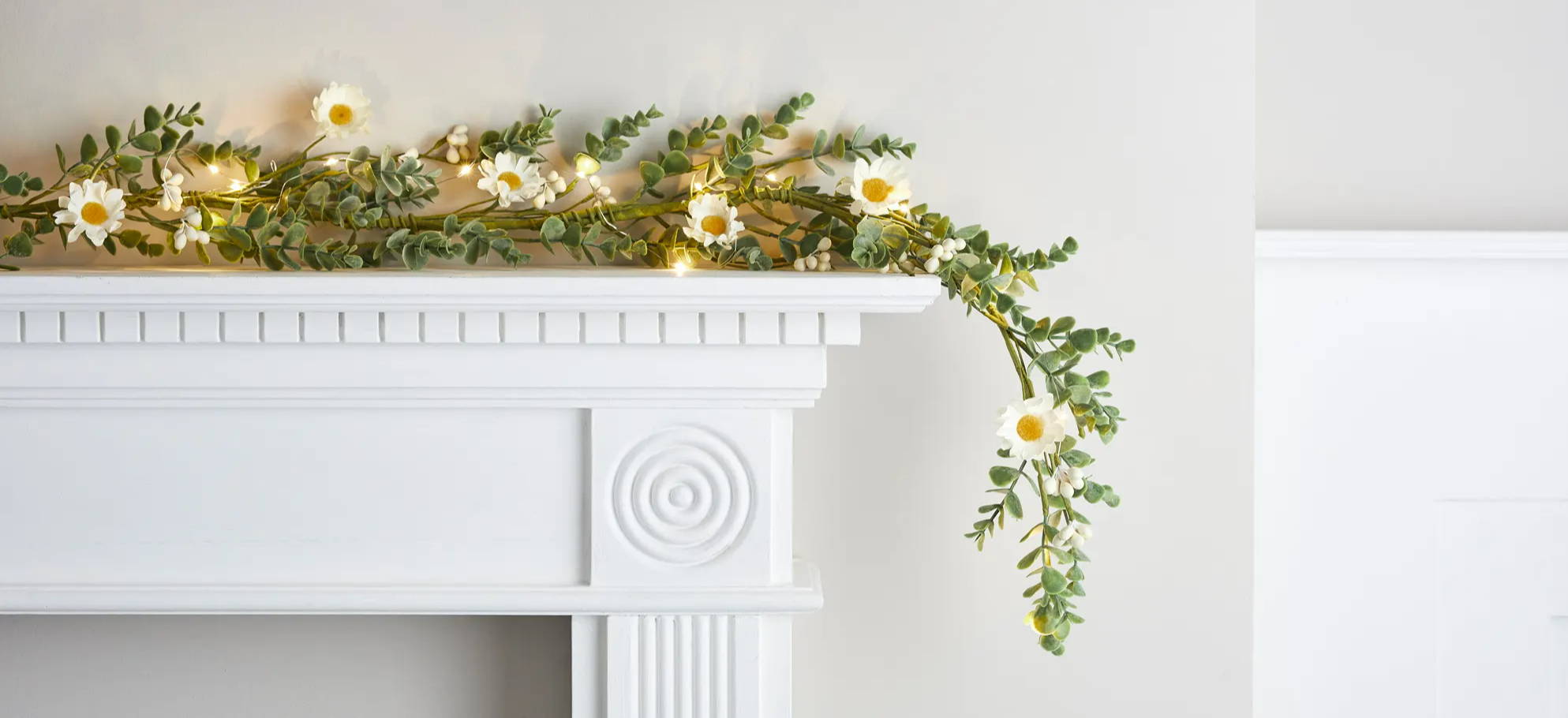 Daisy and foliage artificial garlane with warm white micro lights on a white mantlepiece.