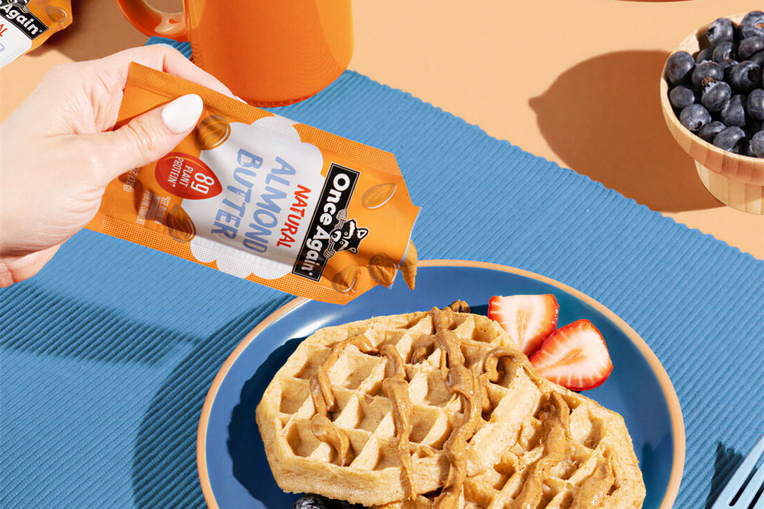 Hand pouring syrup over a waffle with strawberries and blueberries on the side.