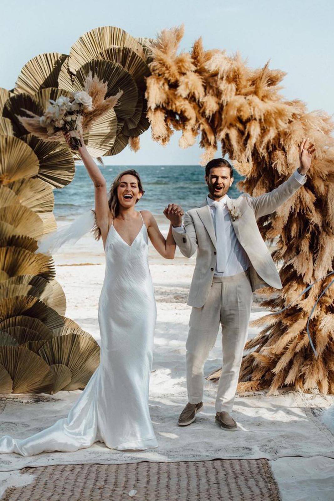The bride and groom stand hand-in-hand with the breathtaking turquoise waters of the ocean in the background, with a stunning floral archway framing the scene.