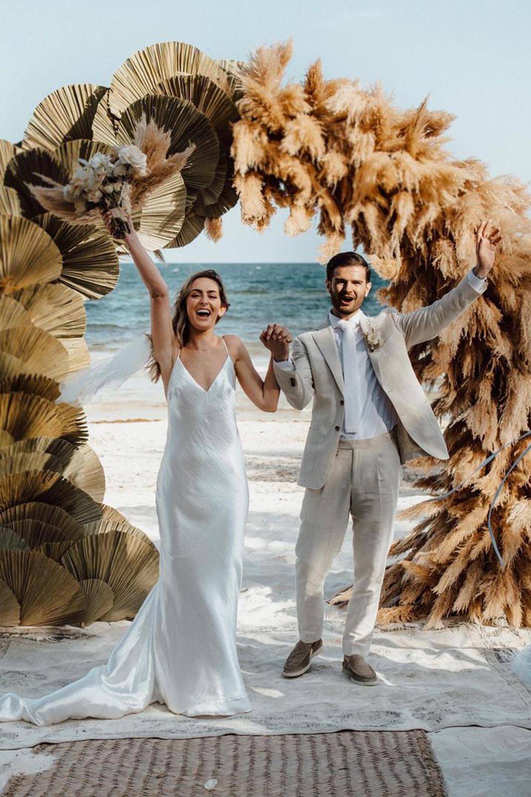 Los novios se cogen de la mano con las impresionantes aguas turquesas del océano de fondo y un impresionante arco floral enmarcando la escena.