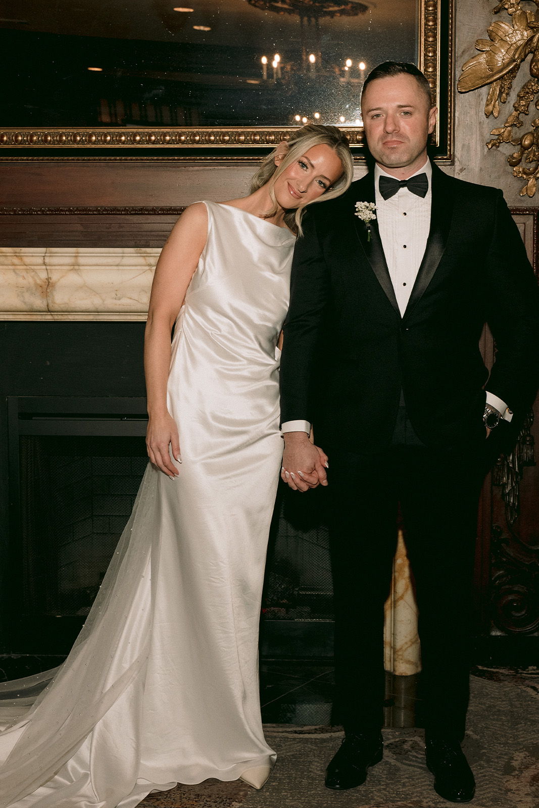 Bride and groom holding hands, posing in formal attire by a fireplace.