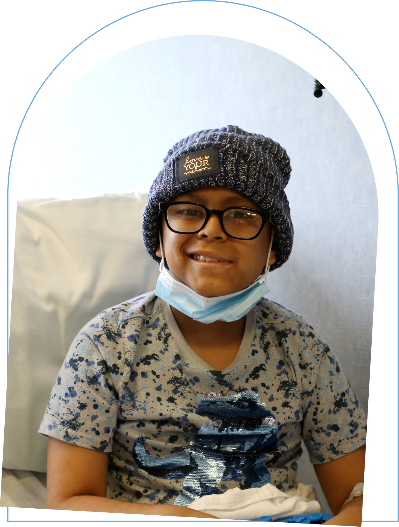 Young boy with mask on seated with a hospital blanket pulled up to his waist wearing a Love Your Melon beanie.