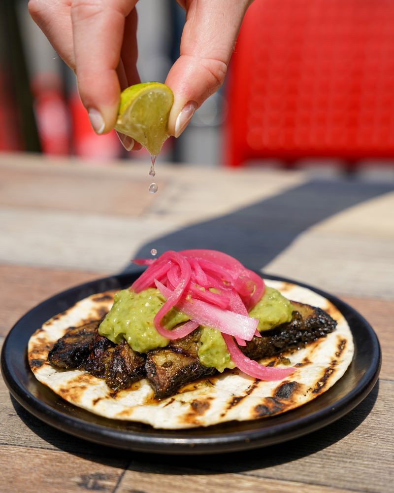 A hand squeezing lime over a taco with meat, guacamole, and pickled onions.