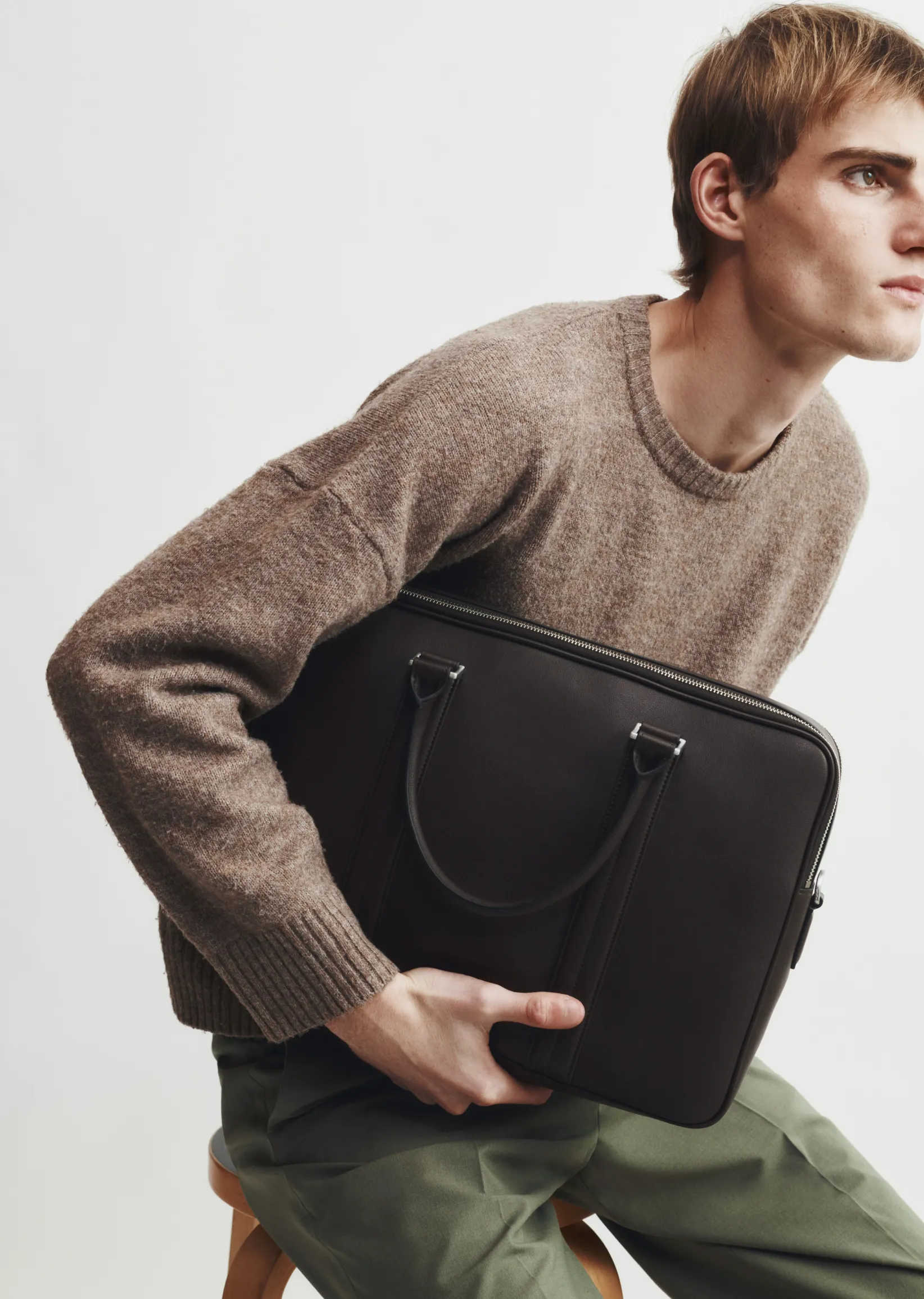A man in a sweater holding a black bag, seated on a stool.