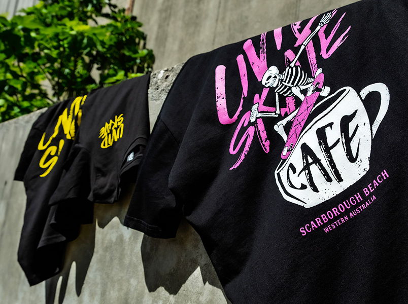 Various black t-shirts screen printed with the bold pink and yellow Livid Skate Café graphic hanging on an industrial cement wall