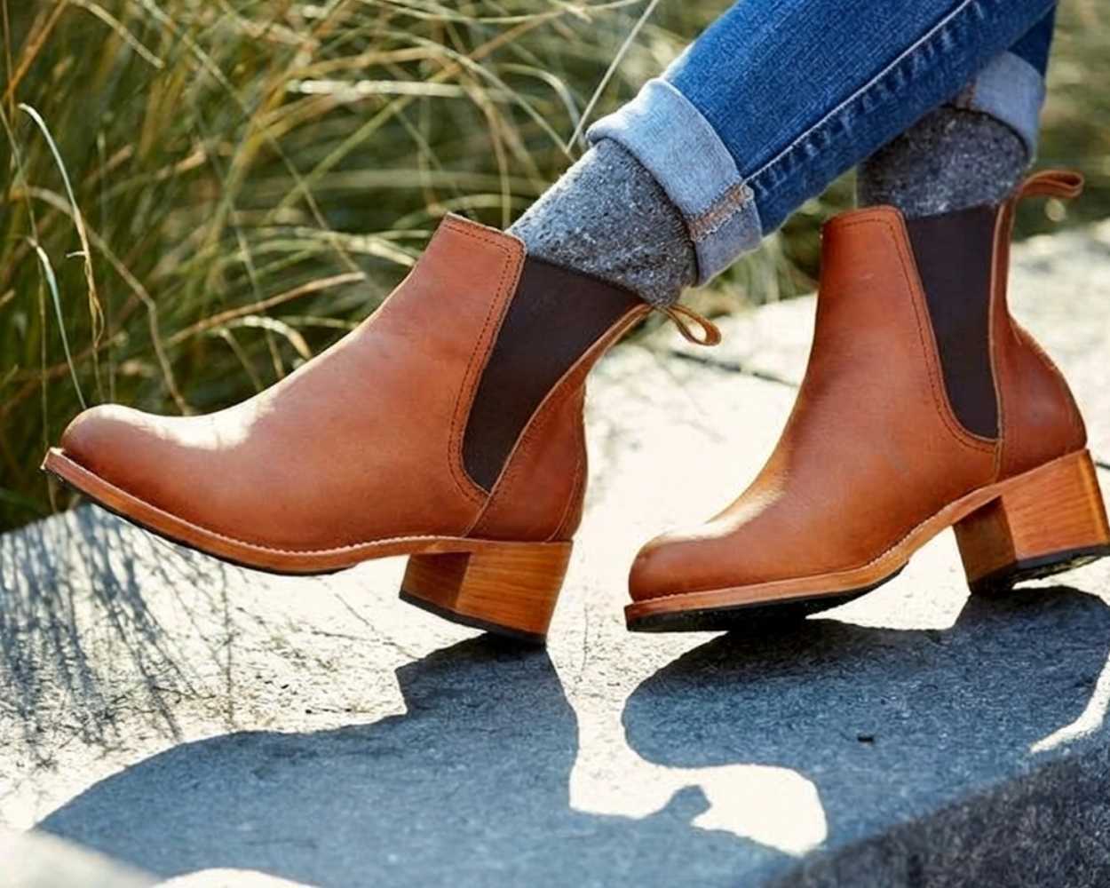 An outdoor lifestyle shot from the calves down of a person sitting on a stone ledge with tall grass in the background. They are wearing cuffed blue jeans, thick grey marl knit socks, and a pair of light brown or caramel leather heeled Chelsea boots with dark brown elastic side panels, a rear pull tab, and a substantial stacked leather heel.