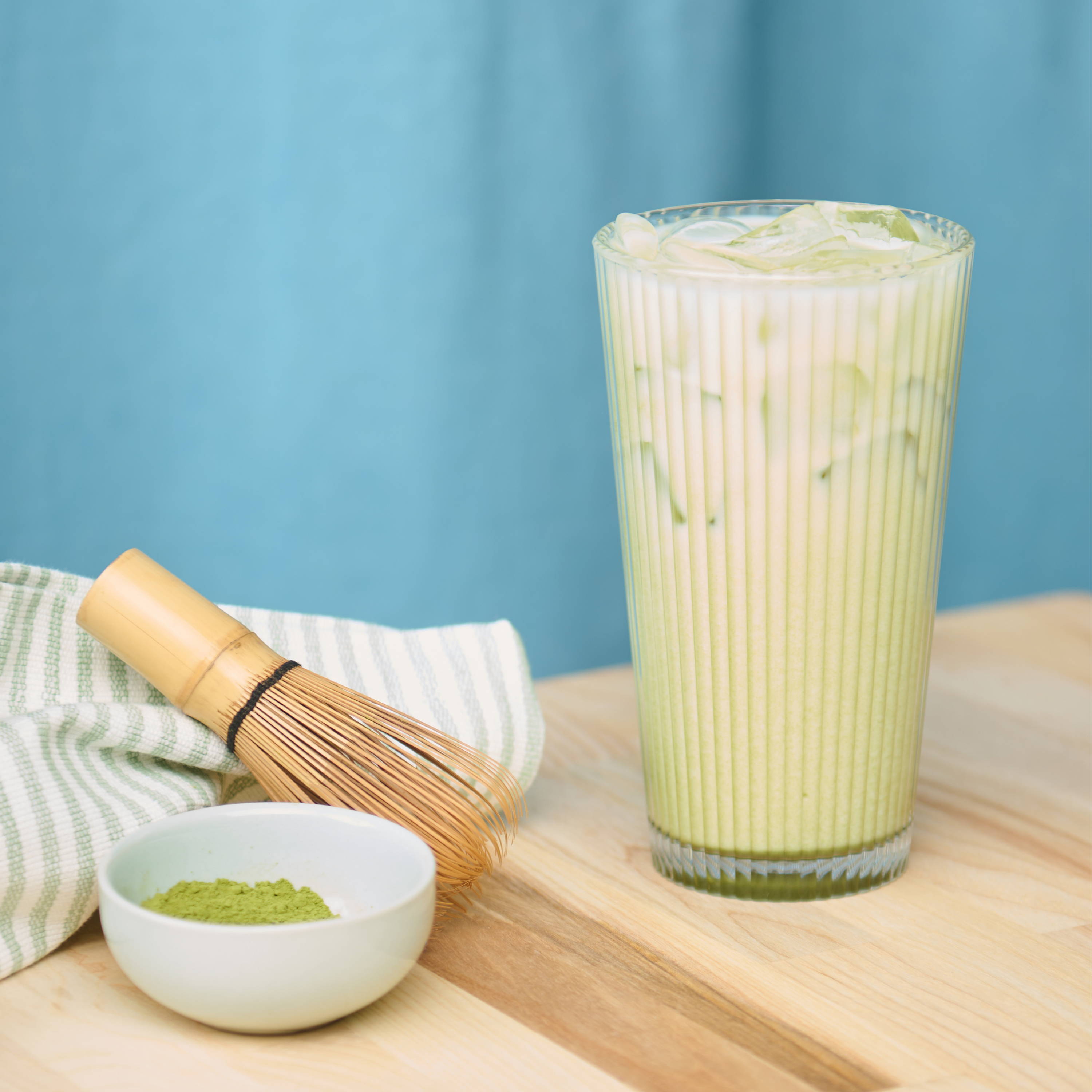 Iced vanilla matcha latte in glass beside a bowl of matcha and whisk