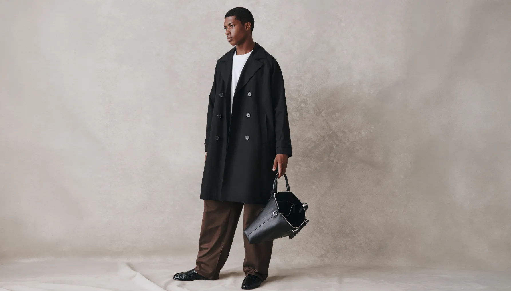 Person in a black coat holding a bag against a neutral background.