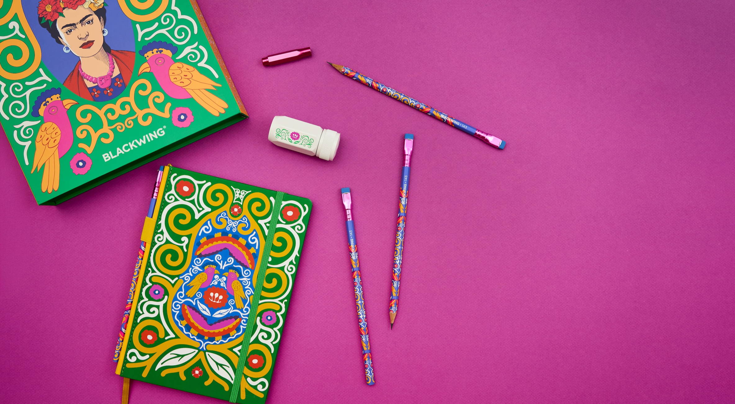 Flat lay of Blackwing Volume 143 gift set, a tribute to Frida Kahlo, featuring a green notebook with Frida's artwork in yellow, white, blue, pink, and red with matching pencils, a sharpener, and a point guard