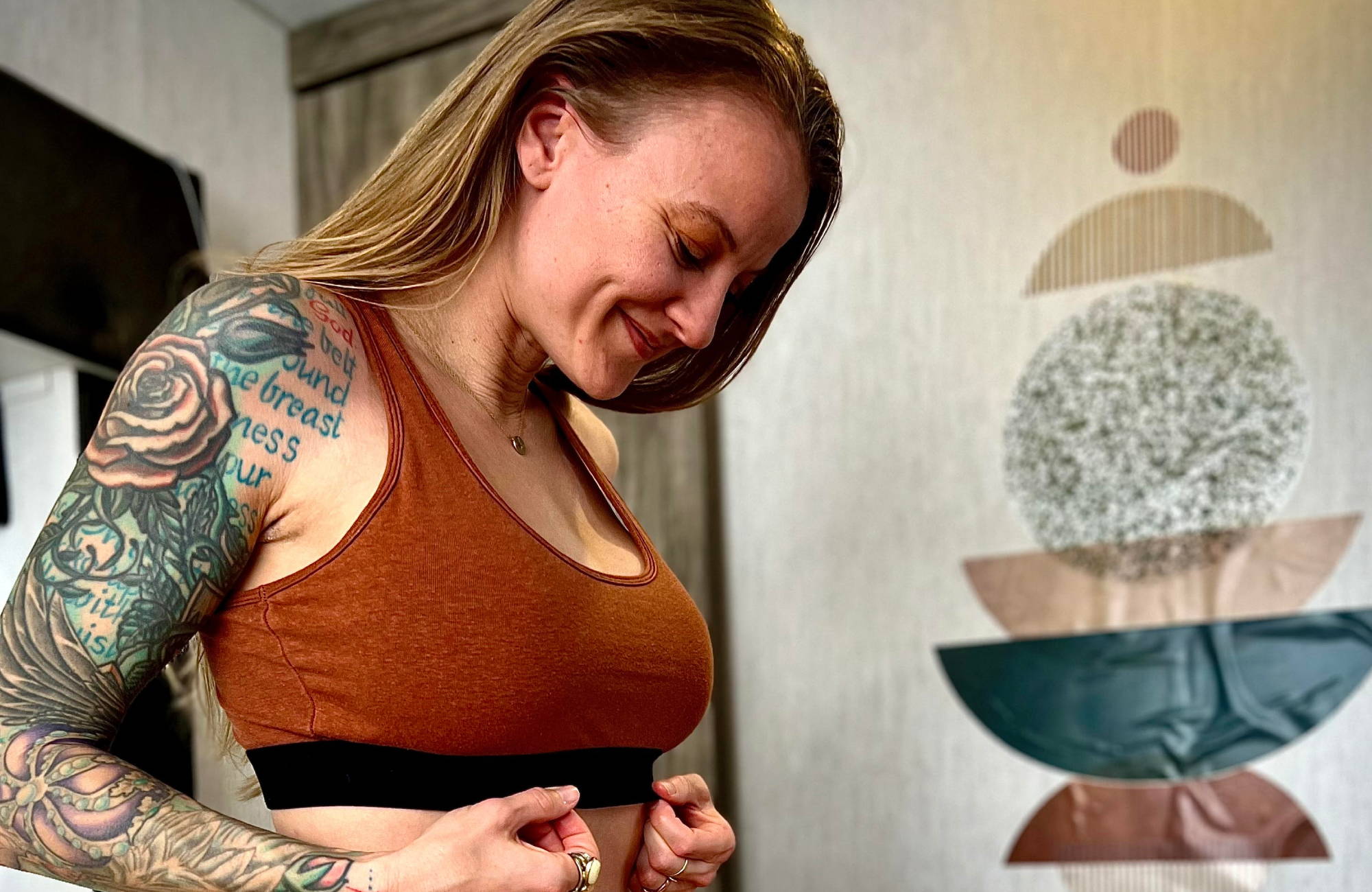 A tattooed woman wearing a brown bralette stands with her hands on her ribs and smiles.