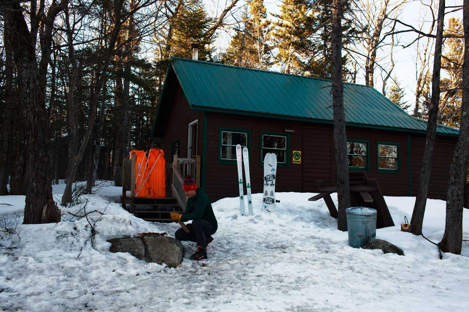 A wooden cabin surrounded by snow and trees, with a person outside.