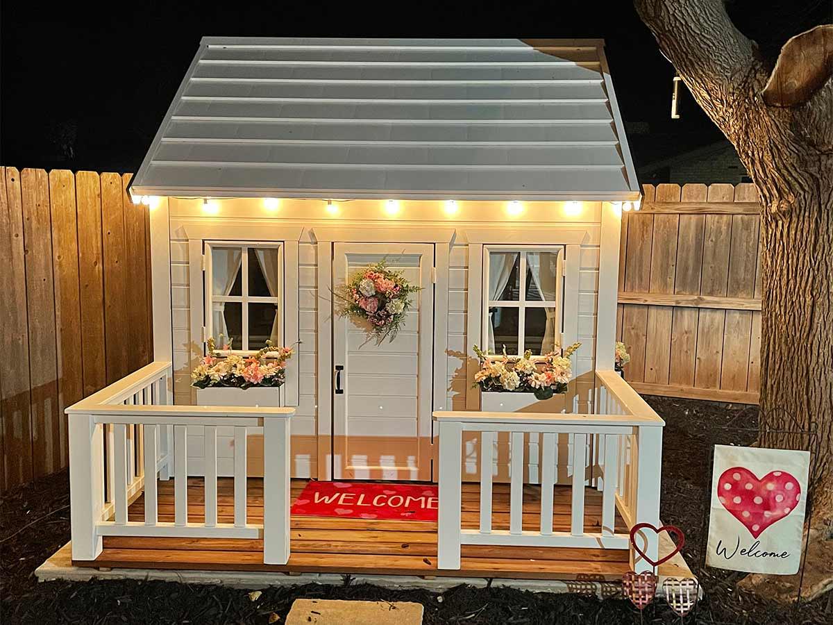 Wooden Playhouse with white roof with lights, flower boxes and a wooden terrace with Valentines Decoration in a backyard by WholeWoodPlayhouses
