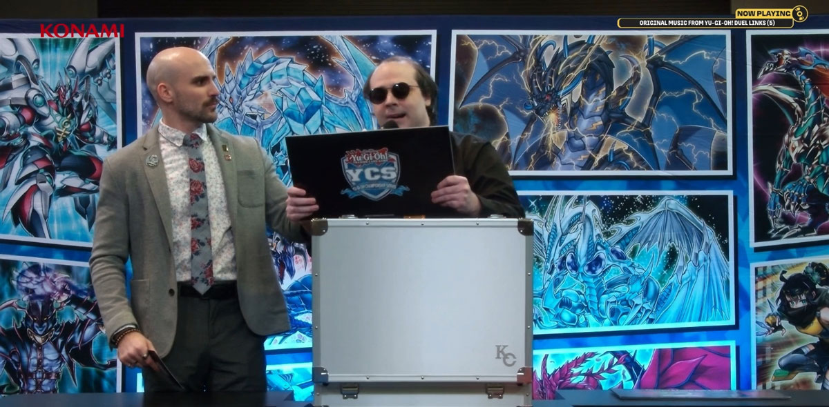 Two presenters on stage with colorful artwork in the background.