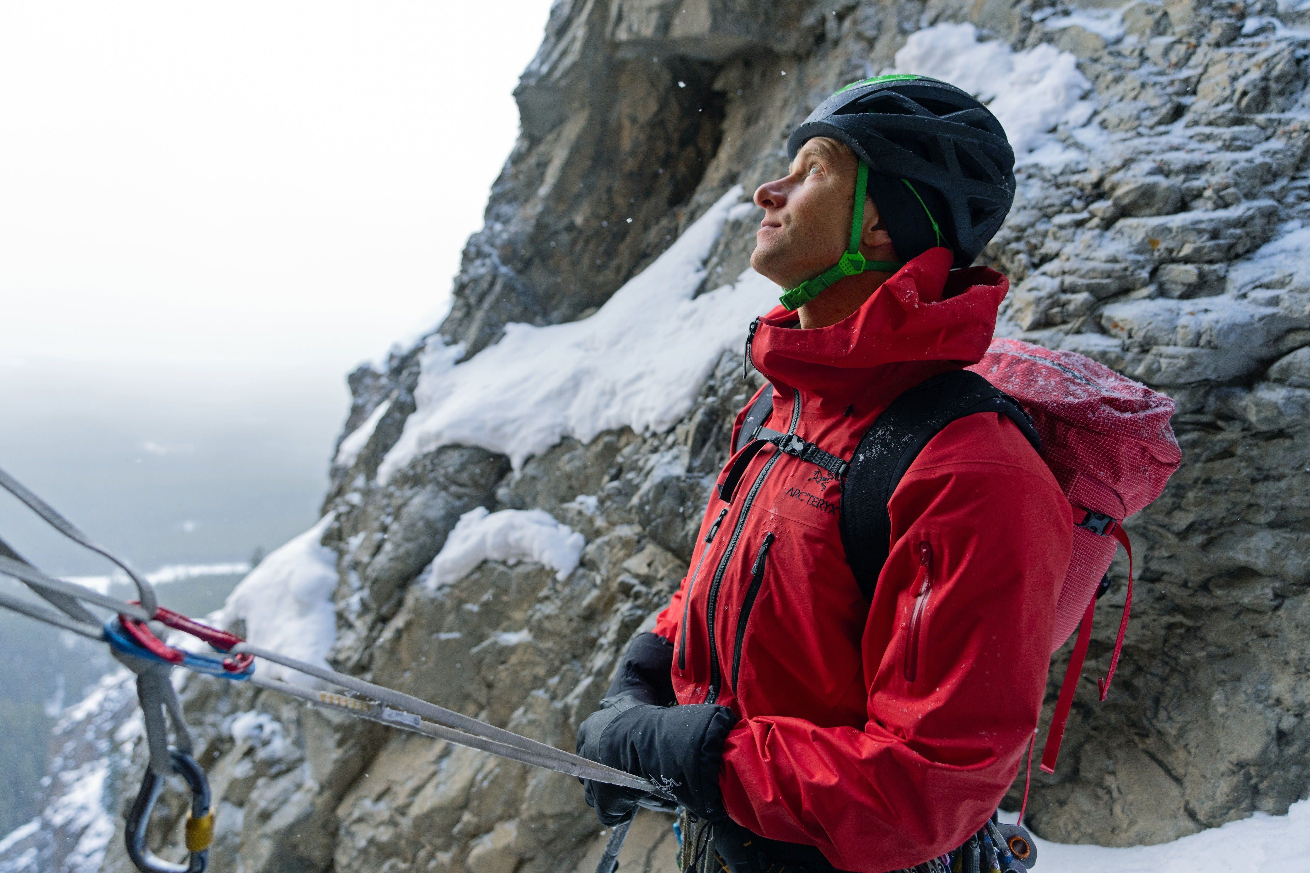 Climber in red jacket resting against a snowy rock face.