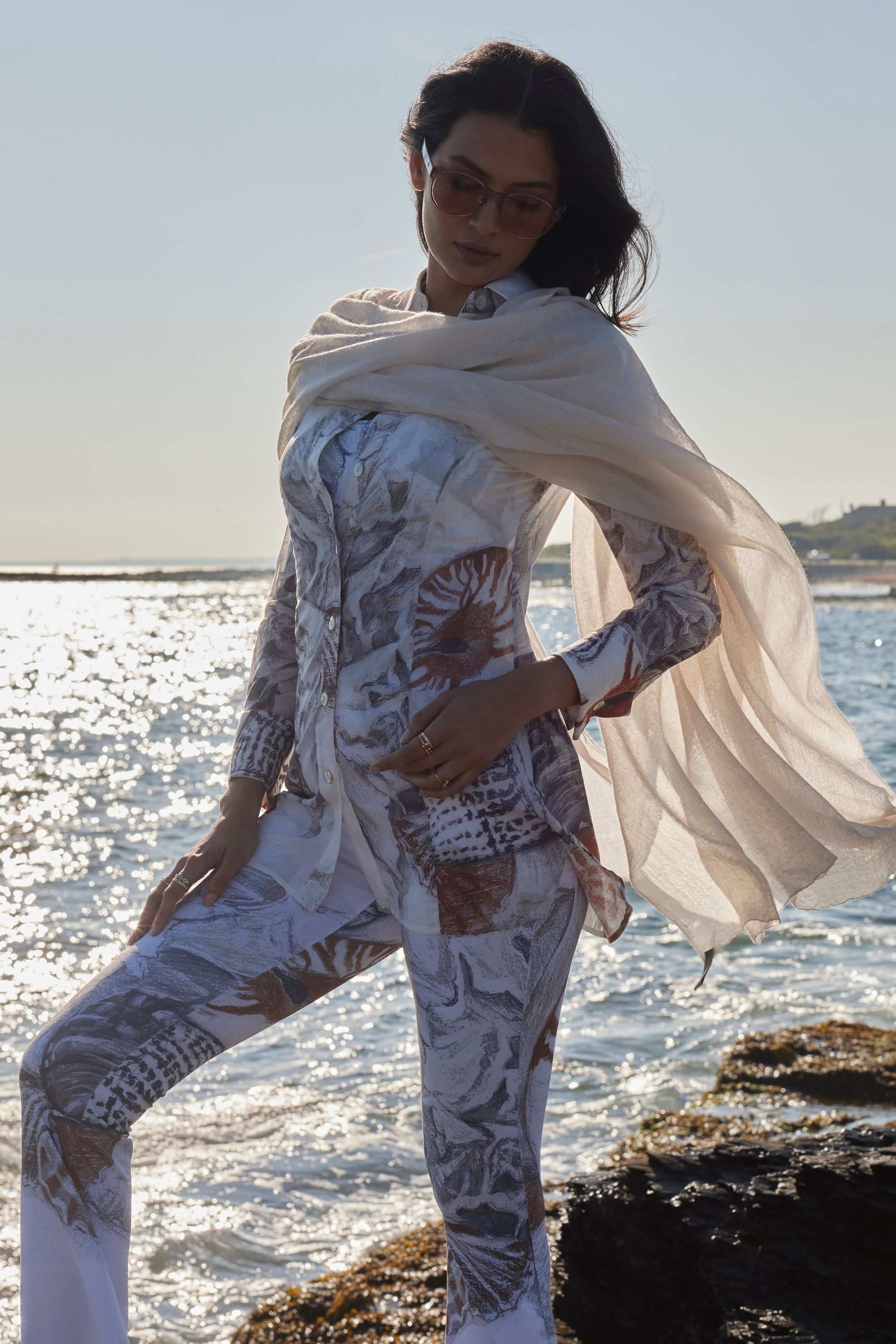 A woman poses in stylish attire by the water, with a scenic background.