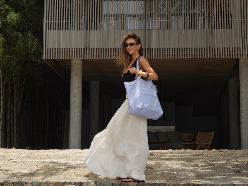 Woman in a flowing white dress, holding a blue bag, standing outdoors.