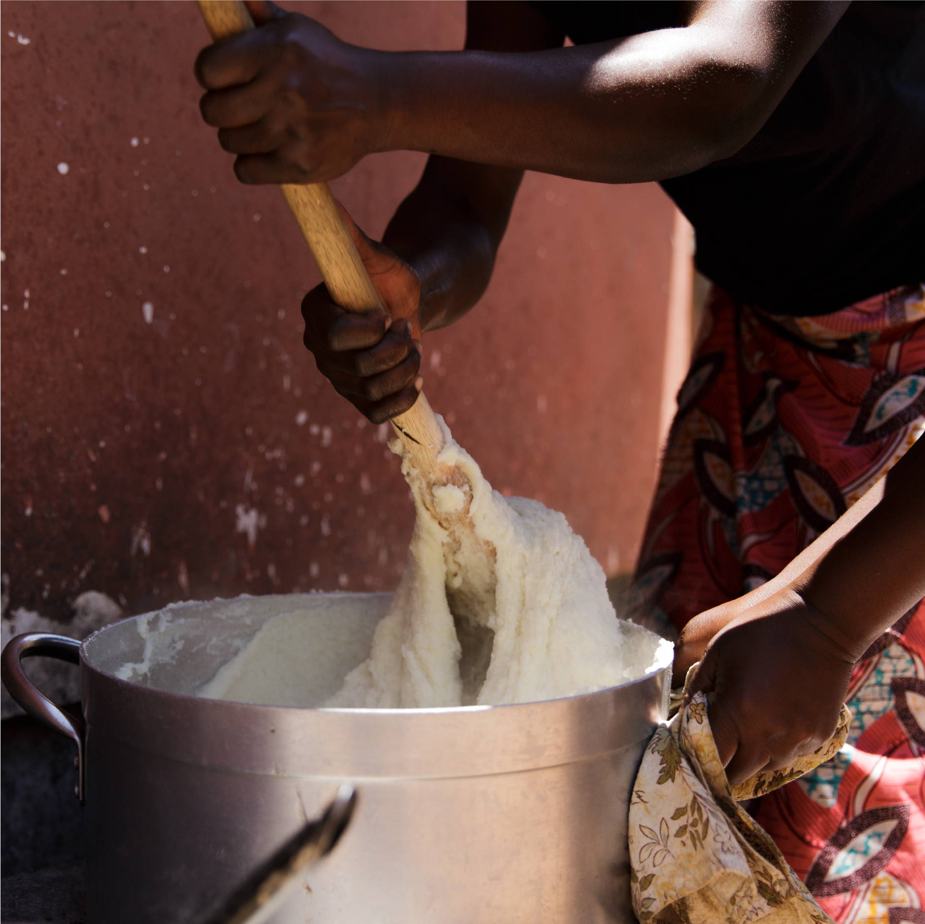 A Zambian women wearing a red skirt stands outside stirring a large stainless steel pot of nshima. 