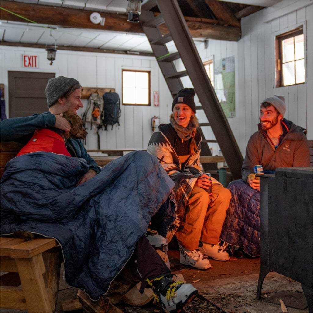 Three people sitting with blankets around a wood stove fireplace