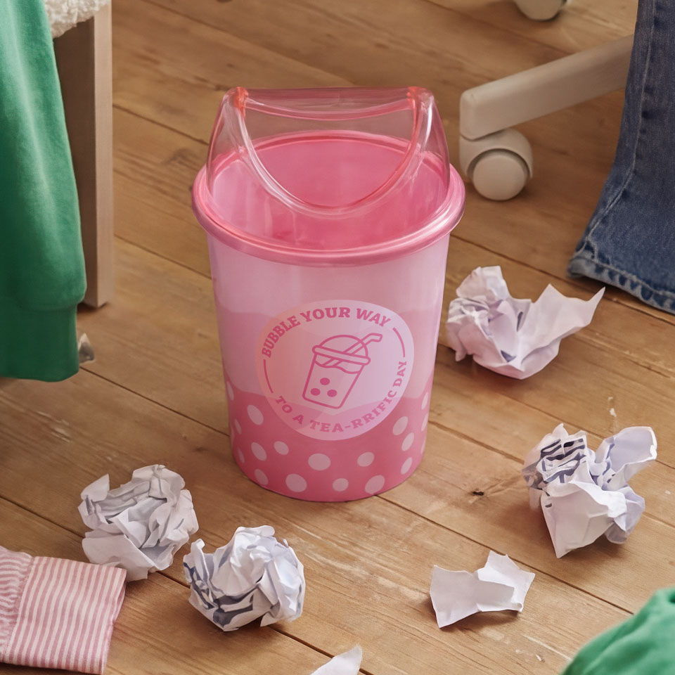 A pink wastebasket surrounded by crumpled paper on a wooden floor.