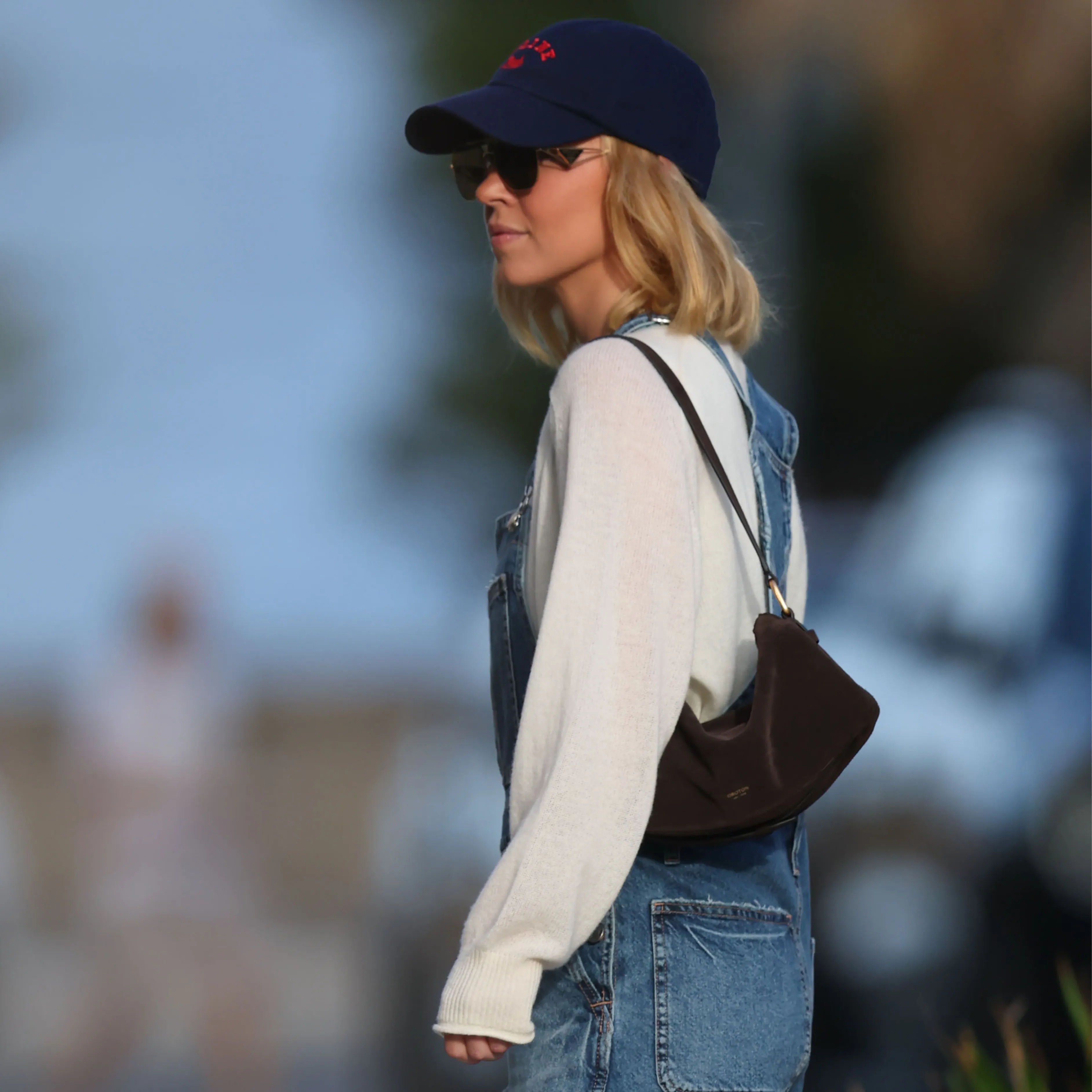 Woman in a cap and sunglasses wearing overalls, walking outdoors.
