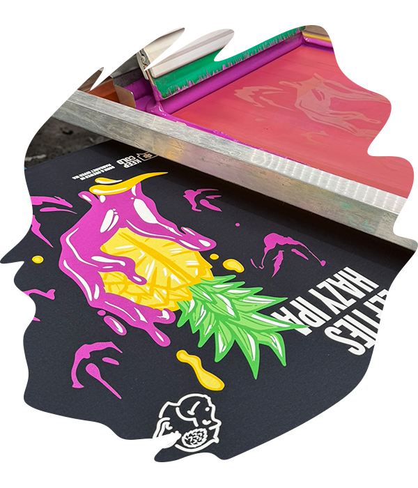 Screen printing squeegee applying bright pink, yellow and green spot colour inks onto black t-shirt with pineapple graphic design