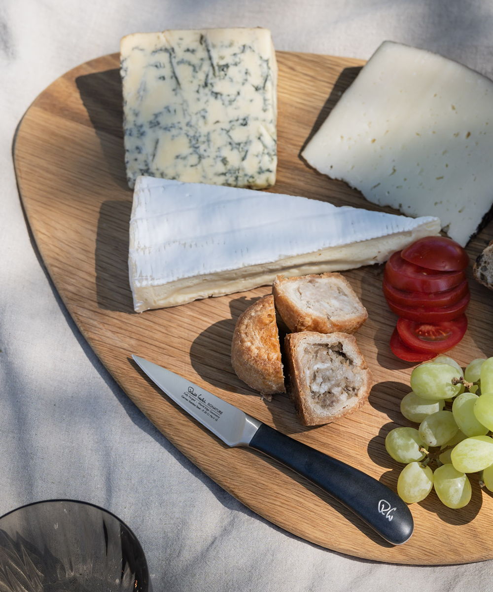 A wooden board with various cheeses, grapes, tomatoes, and a paring knife.