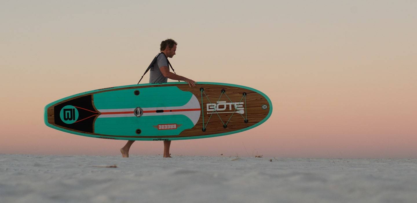Man carrying his Breeze 10′6″ Classic Teak Paddle Board on the beach using a Travelink Sling