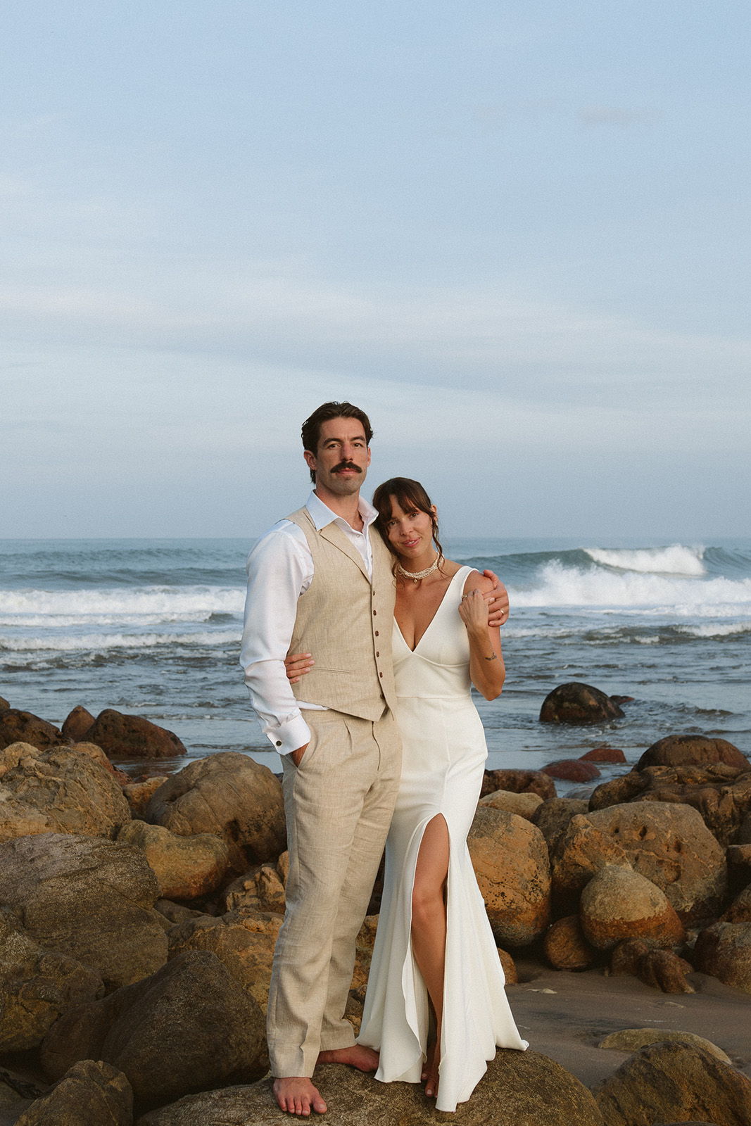 Couple in wedding attire by the ocean on rocky shore.
