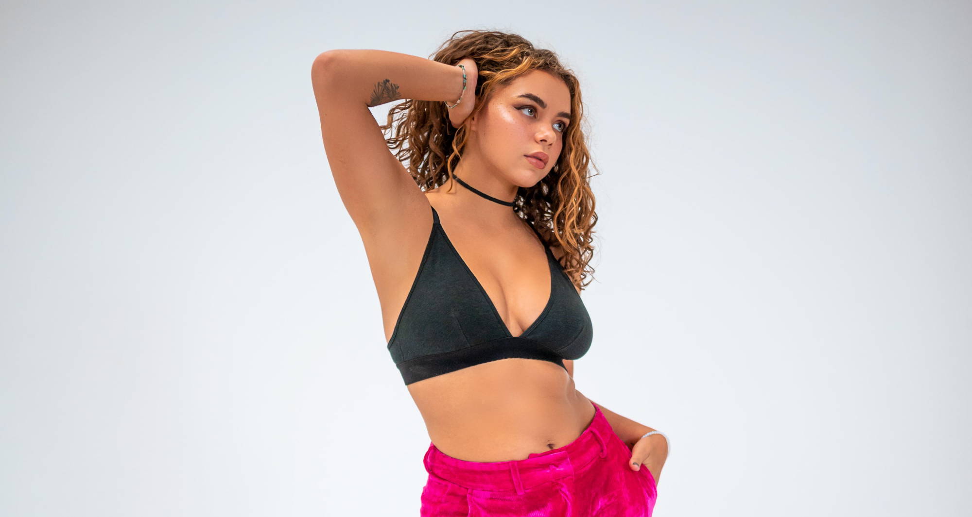 A woman with curly hair wearing a black bralette and pink pants poses in front of a white background