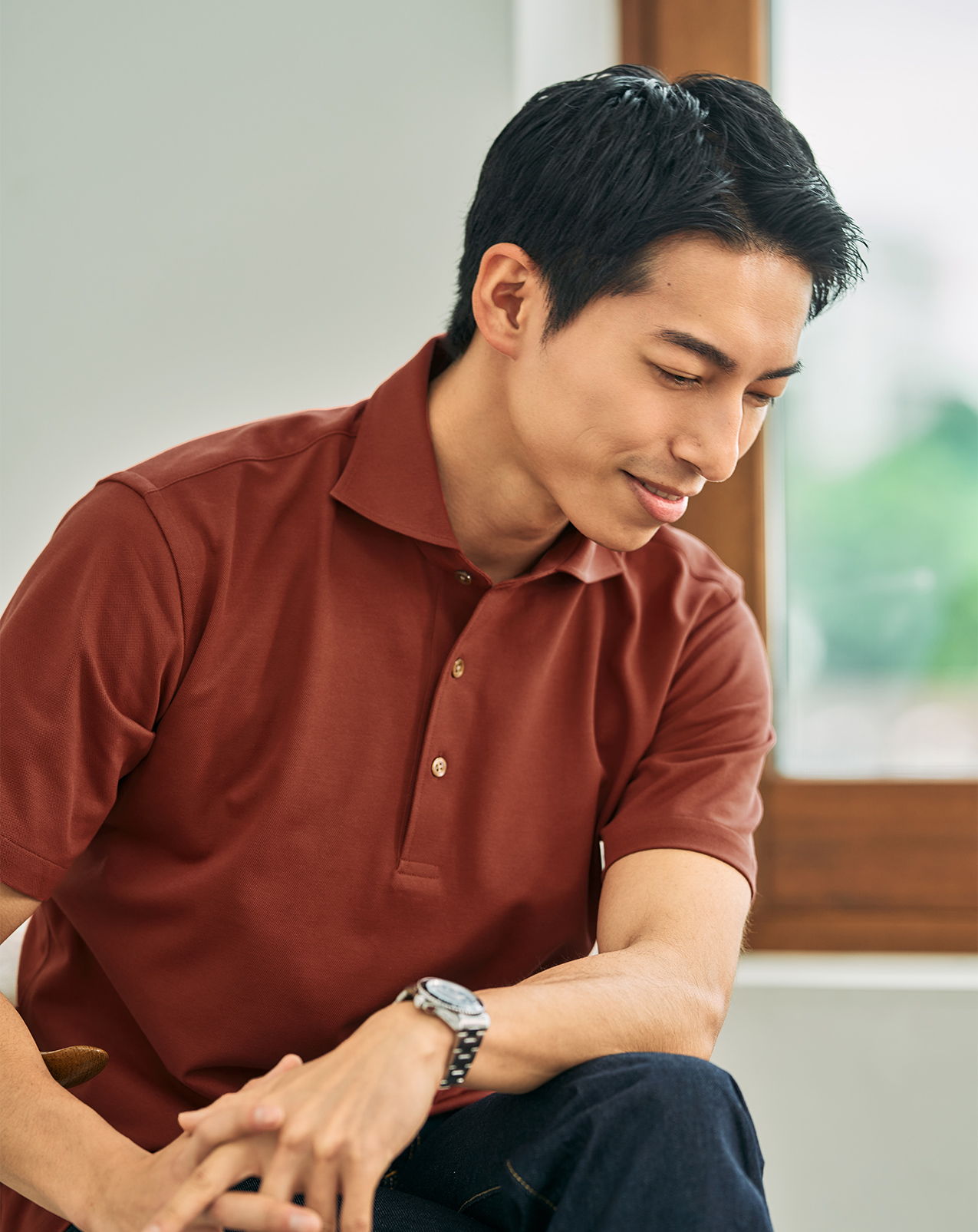 Man in a brown polo shirt looking down thoughtfully by a window.