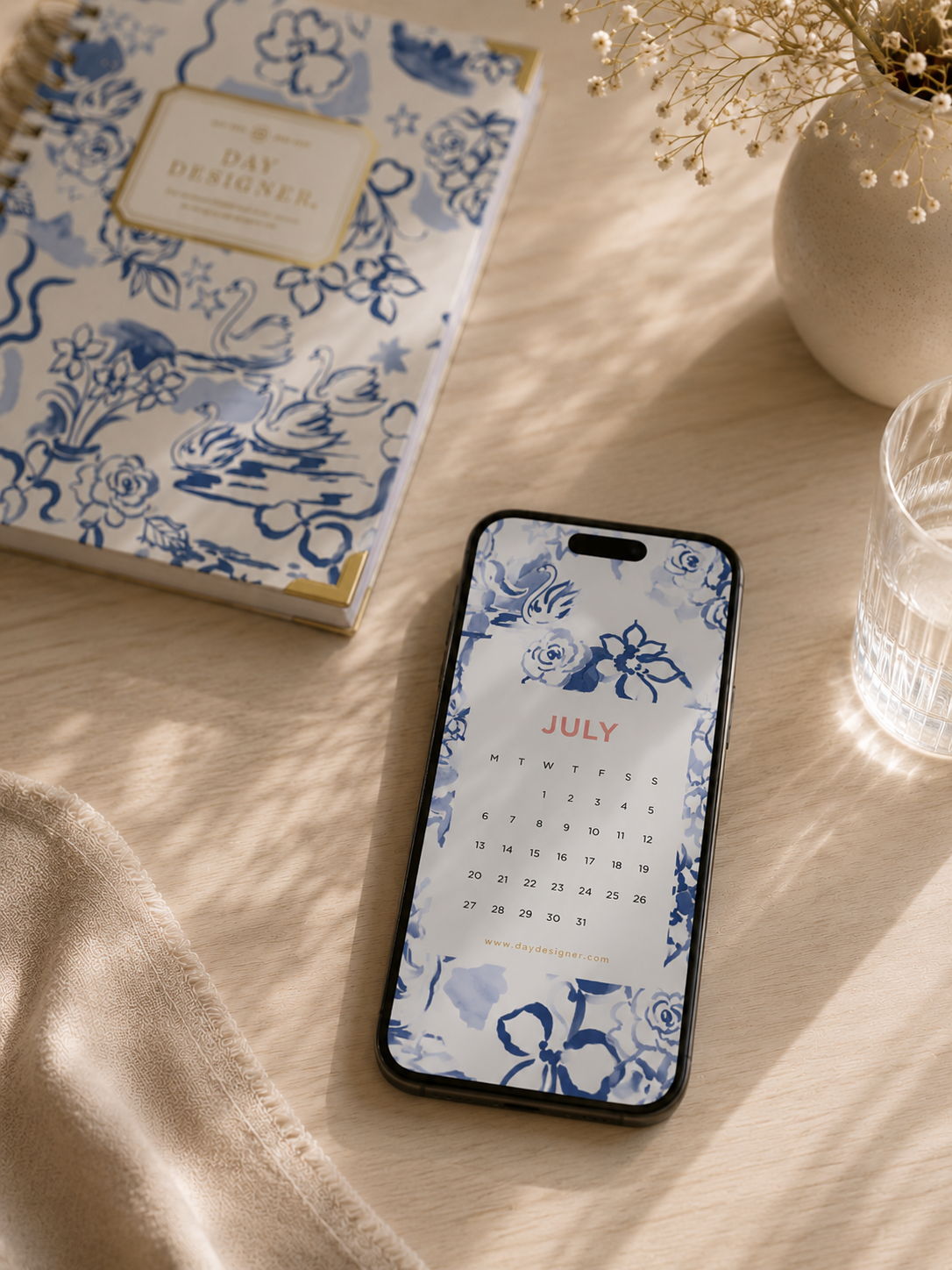 mobile phone with swan pattern july digital calendar, matching day designer planner on beige tablecloth, water glass, vase with flowers