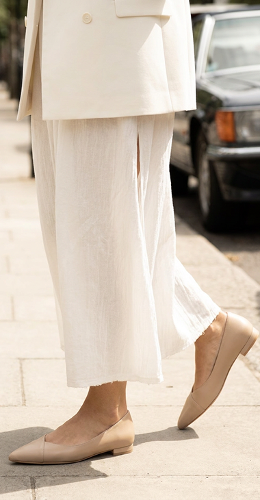 A person wearing white pants and beige flats walking on a sidewalk.