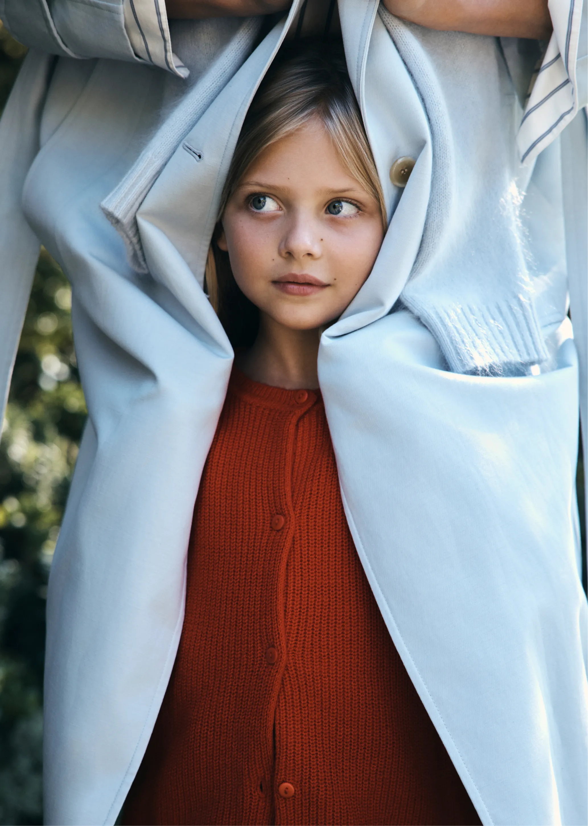 Child peeking out from under a large coat, wearing a red cardigan.