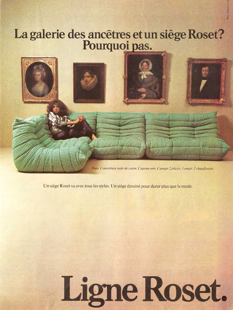 A person sitting on a green couch Togo Sofa with framed portraits on the wall.