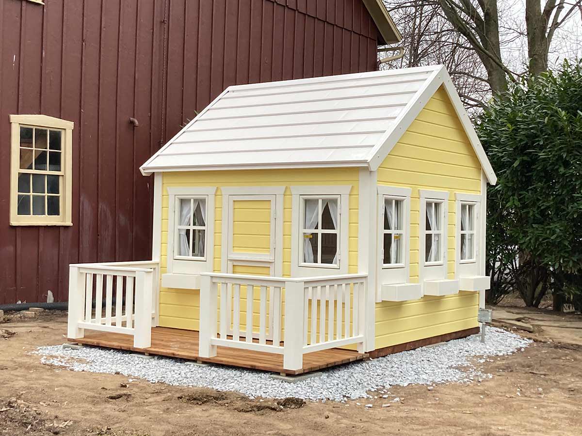 Yellow custom playhouse with white roof on gravel next to brown building by WoleWoodPlayhouses