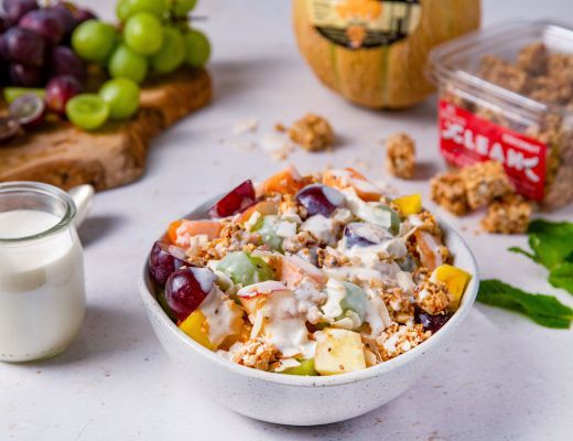 Bowl of mixed fruit and granola with yogurt, surrounded by grapes and pumpkin.