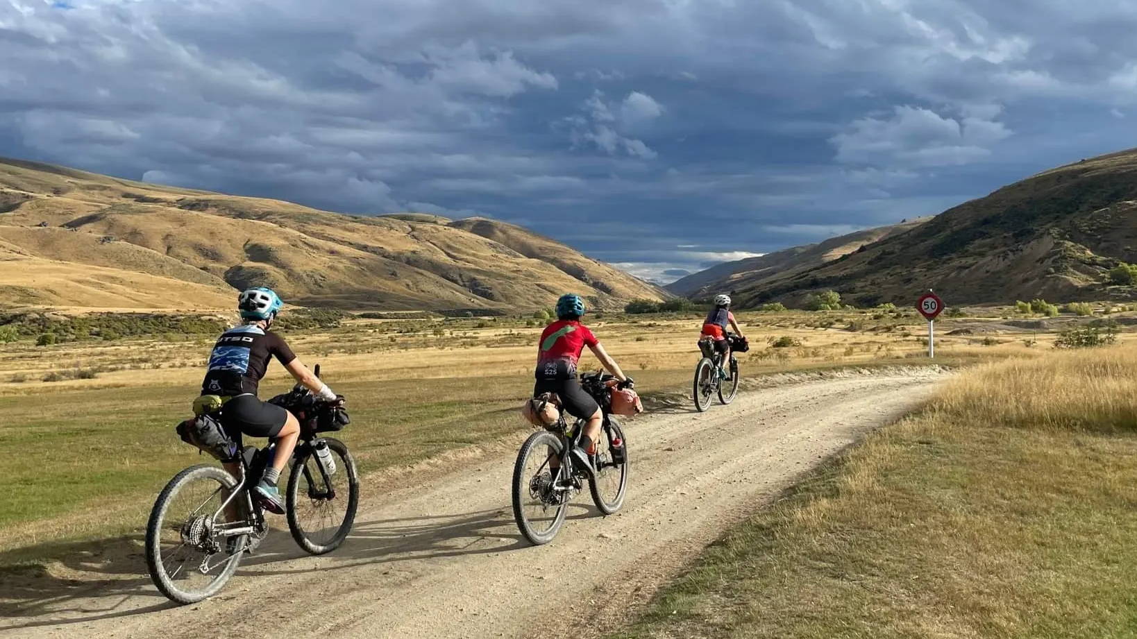 Three cyclists ride along a gravel road while dark storm clouds swirl over the hills in the distance.