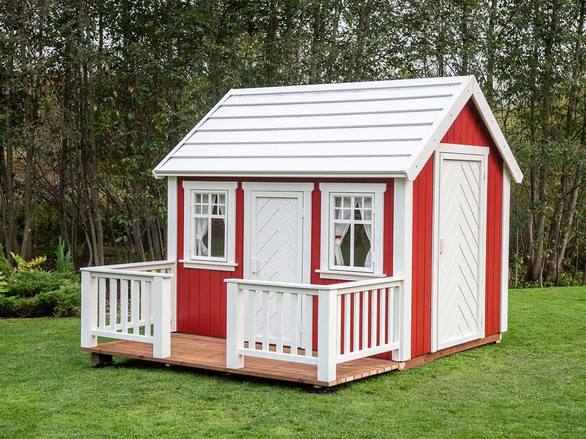 Wooden Playhouse in red and white color with a wooden terrace and white roof on green lawn with forest on the background by WholeWoodPlayhouses