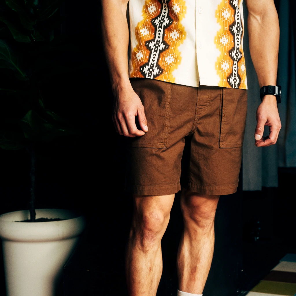 Man wearing a patterned shirt and brown shorts, standing indoors.