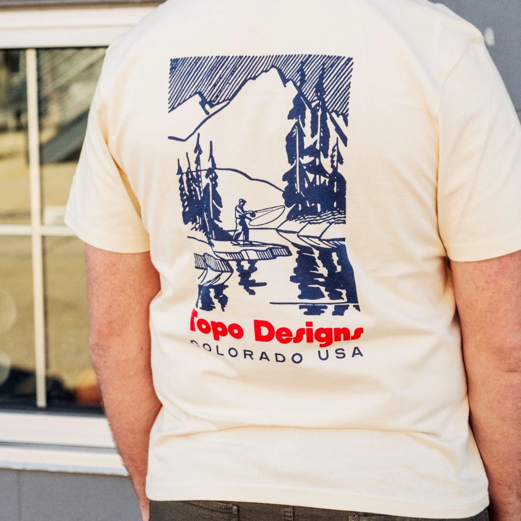 Person wearing a cream t-shirt with mountain and tree graphic design.