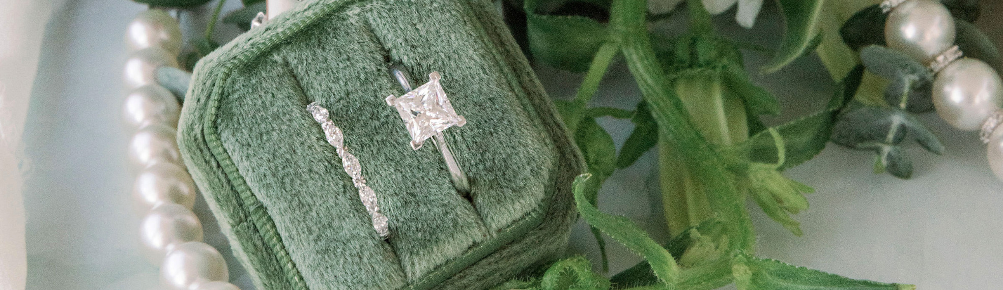 Princess cut solitaire white gold engagement ring with delicate alternating marquise and round diamond band in moss green ring box with green florals and pearls in the background
