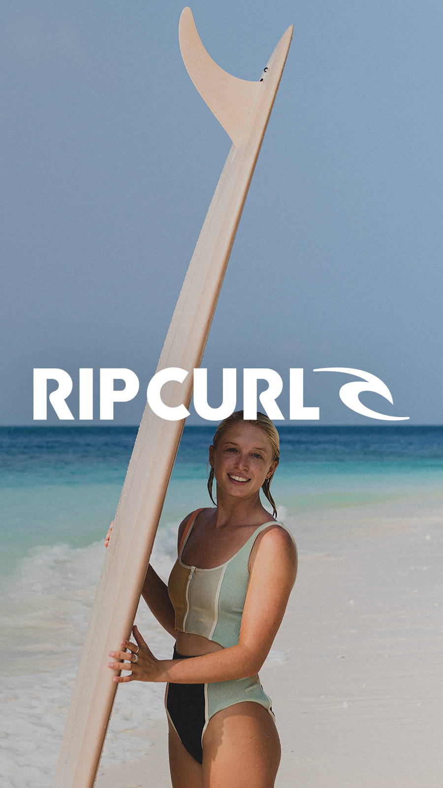 Young woman standing on beach in Rip Curl bathing suit holding up surf board