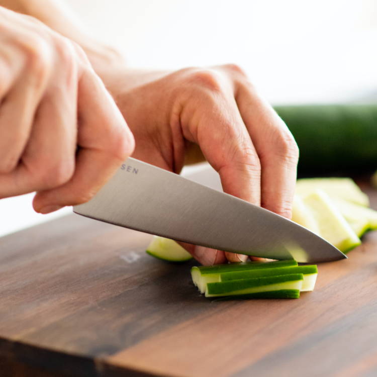 A chef using a Misen Utility Knife to slice cucumber into thin vegetable sticks on a dark wood cutting board.
