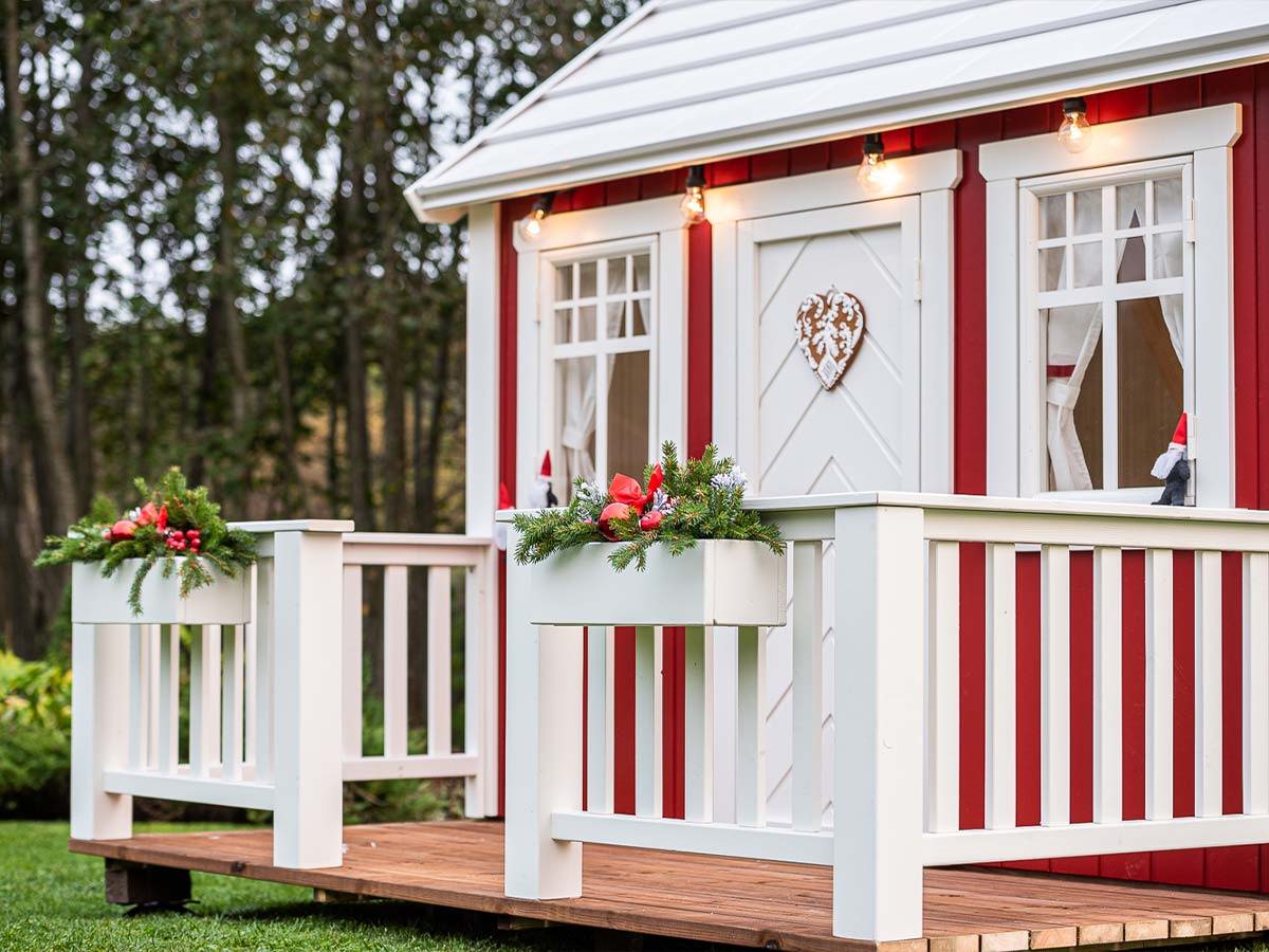 Red and white wooden playhouse with a white porch and Christmas decorations with forest background by WholeWoodPlayhouses