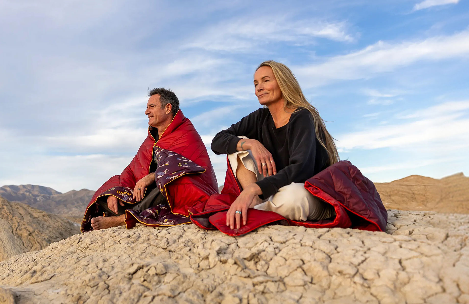 Two people sitting on a rocky surface, dressed in colorful blankets, with mountains behind.
